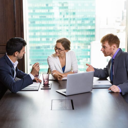 Smiling young businesswoman pointing to laptop screen while sitting at head of table in meeting room. Man sitting on her right is telling something. Man sitting on her left is pointing to the screen.